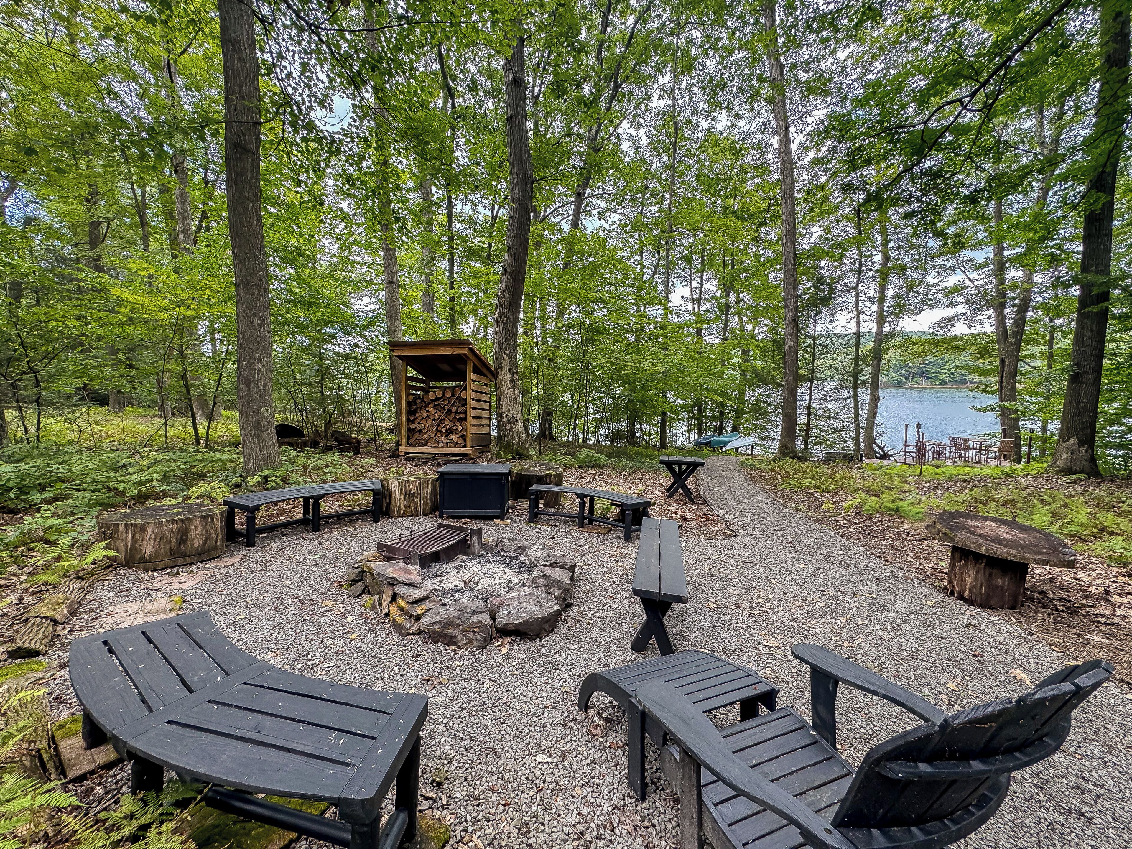 An outdoor seating area with benches and a fire pit, surrounded by trees and a lake view.