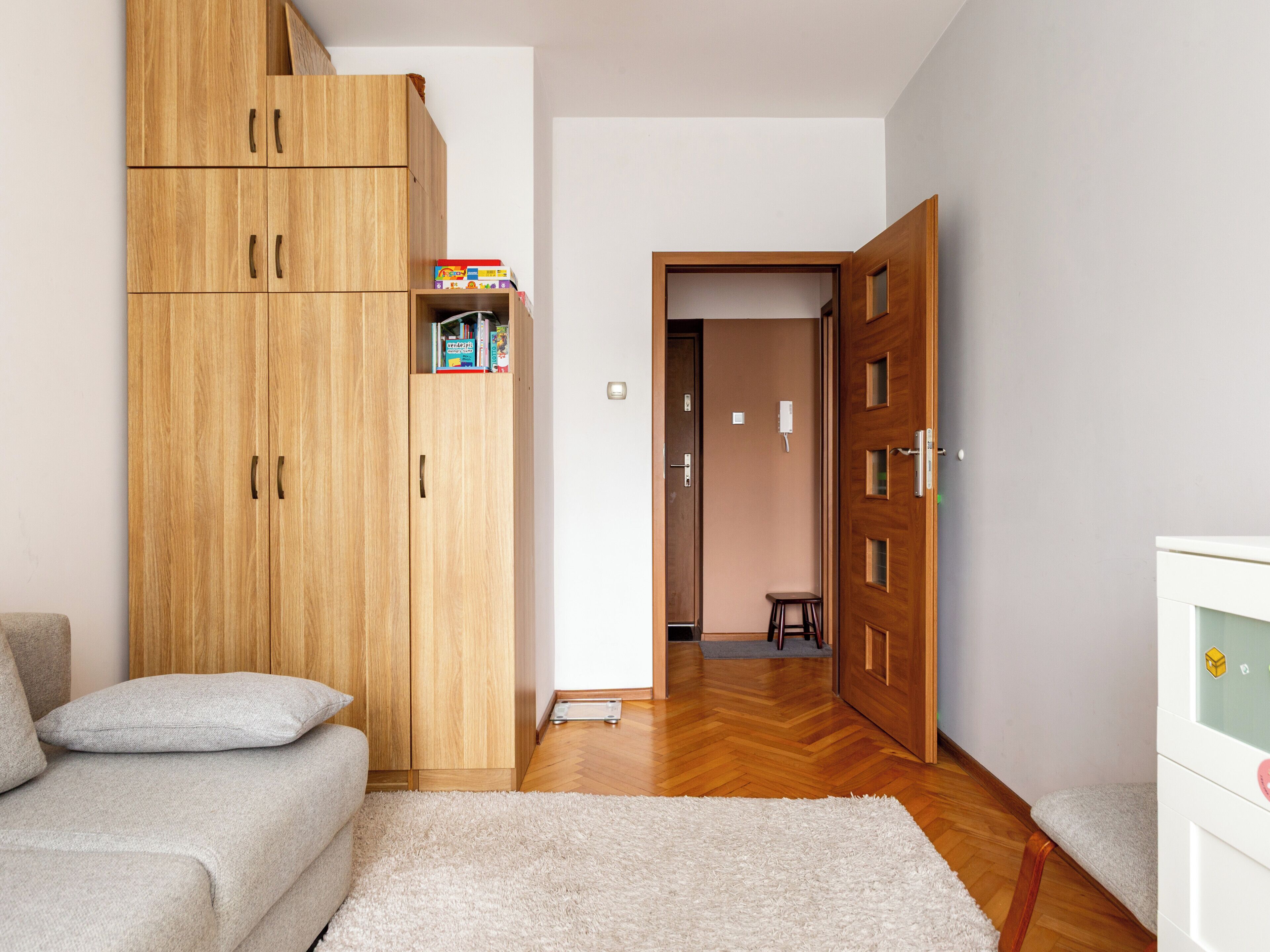 A room with a wooden wardrobe, a sofa, a small stool, and a white cabinet.