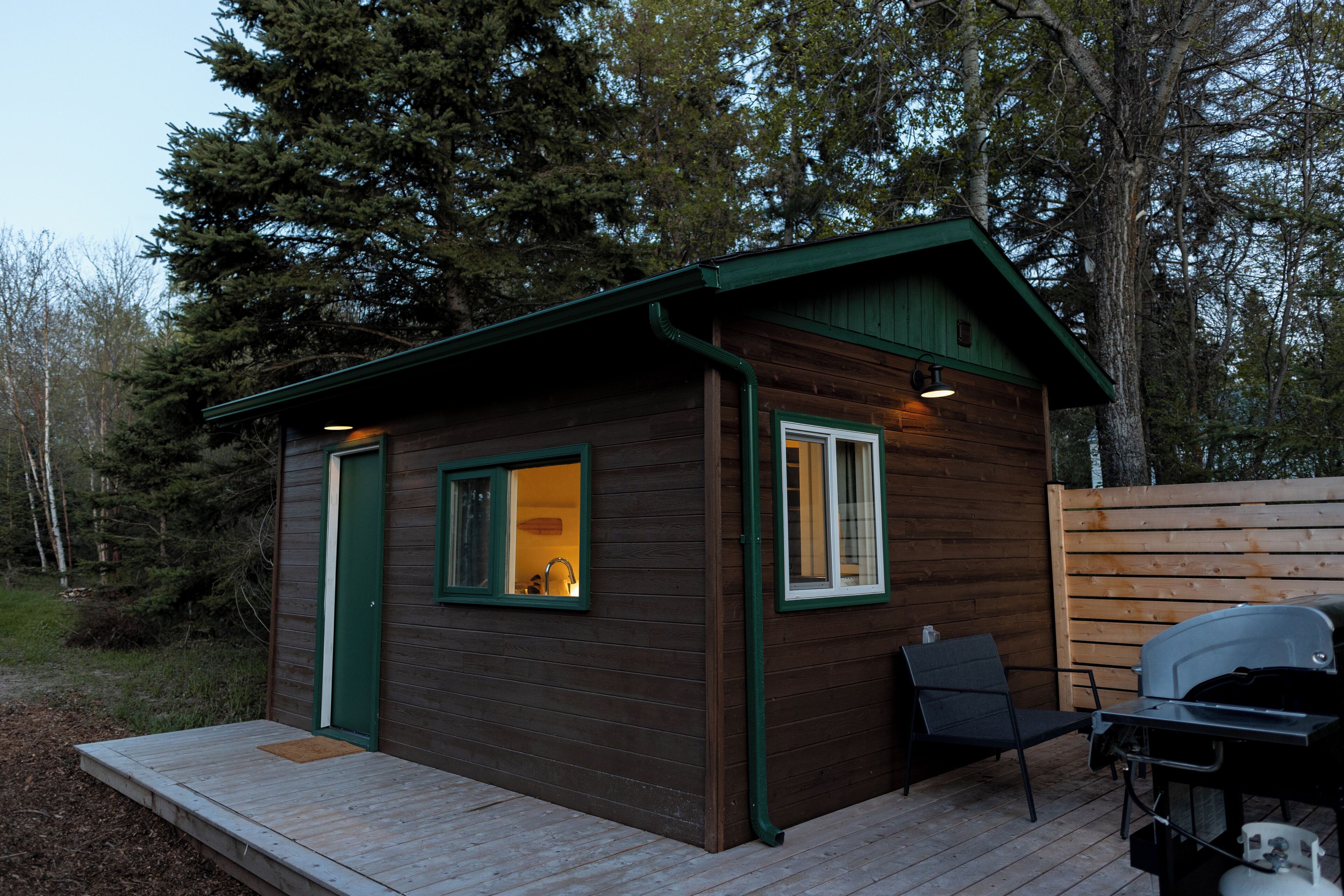 A small wooden cabin with a deck, a grill, and a chair.