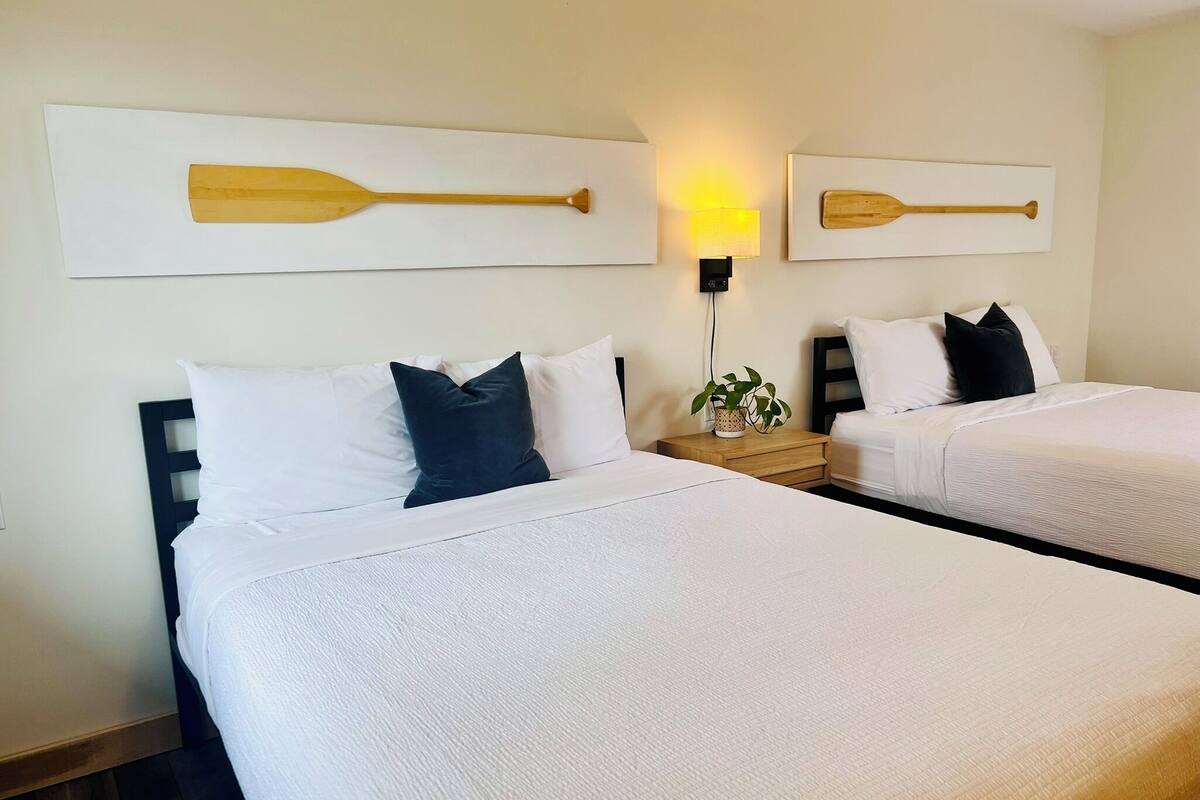 A hotel room with two beds, a nightstand with a lamp, and two wall-mounted paintings of oars.