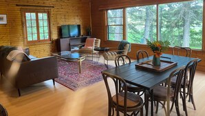 A cozy cabin interior with a wooden ceiling, a TV, a sofa, a dining table with chairs, and large windows offering a view of trees. - Smokey Hollow Motel & Resort (Onanole)
