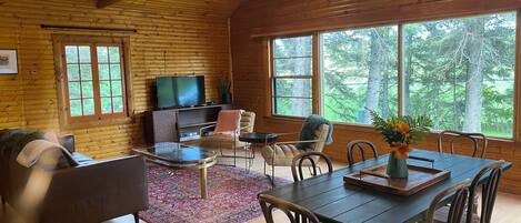 L’intérieur chaleureux d’un chalet avec un plafond en bois, un téléviseur, un canapé, une table à manger avec des chaises et de grandes fenêtres donnant sur la forêt.