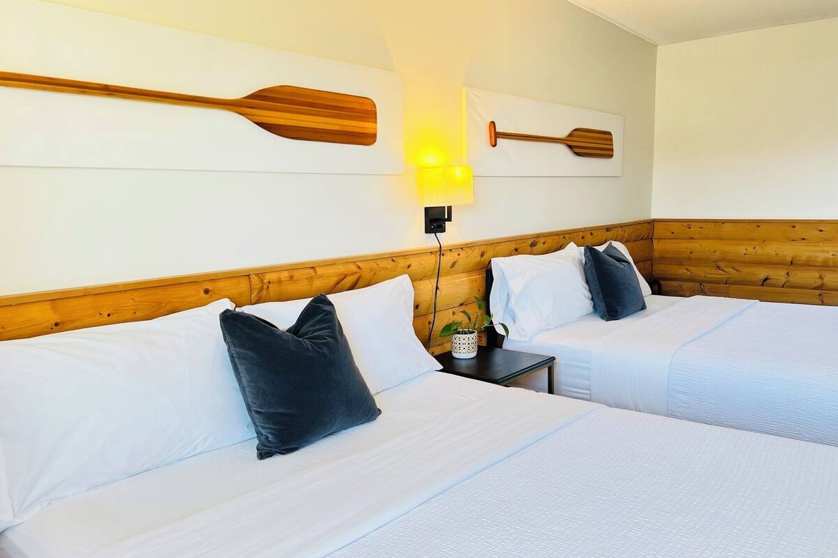 A hotel room with two beds, wooden headboard, white bedding, and wooden paddle wall art.