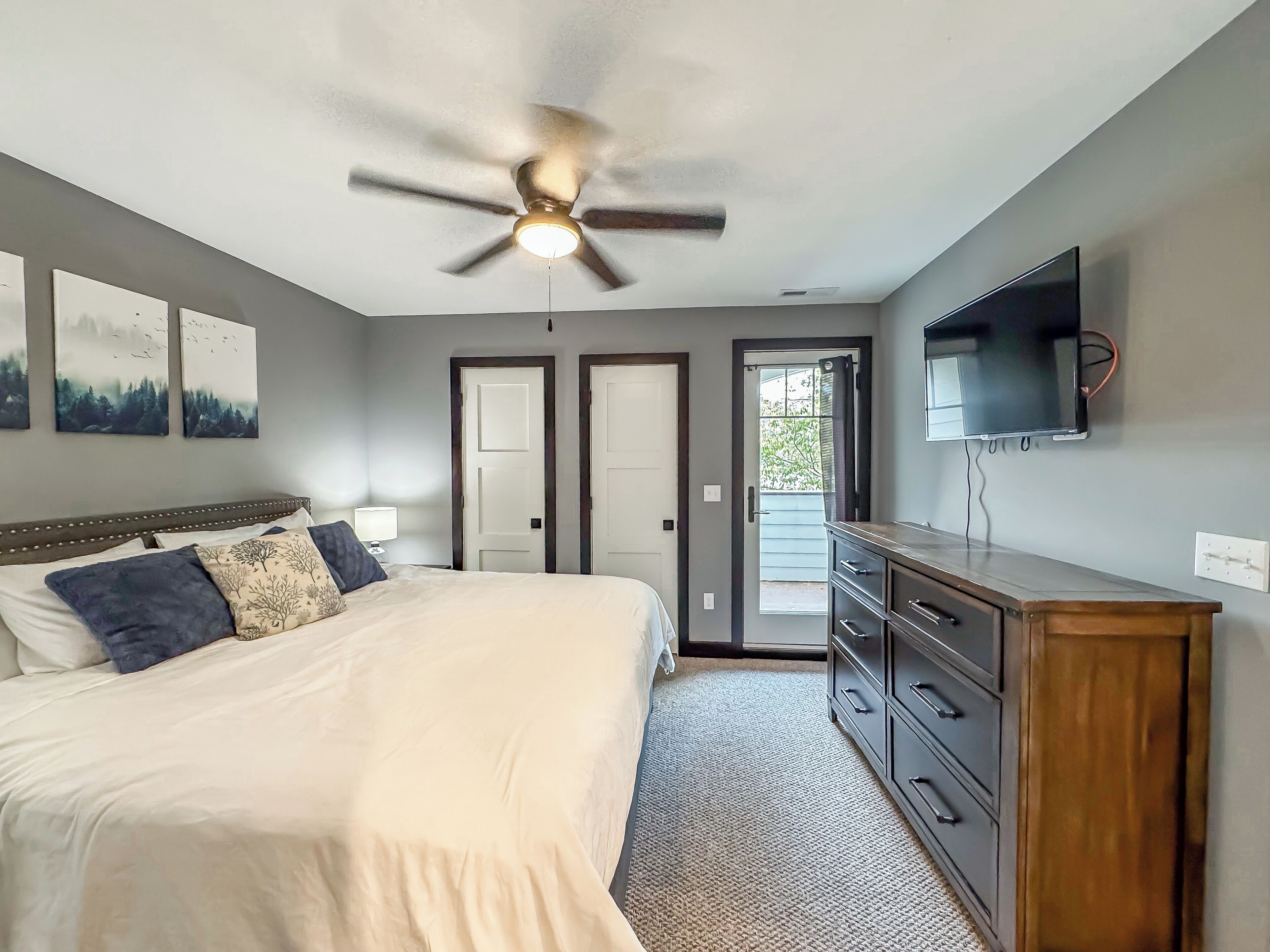 Une chambre à coucher comprenant un lit, un ventilateur de plafond, une commode, un téléviseur et deux portes.