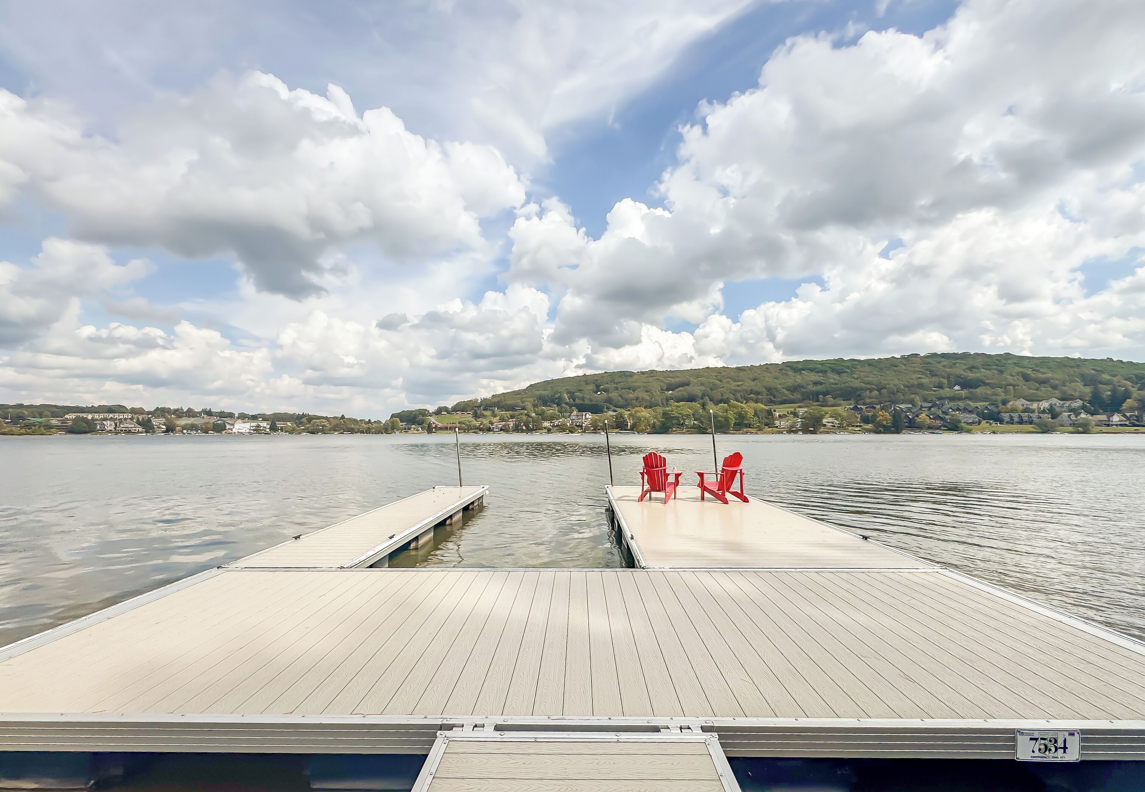Une jetée en bois avec deux chaises rouges, donnant sur un lac paisible, le tout entouré d’un paysage vallonné où l’on aperçoit des maisons au loin.