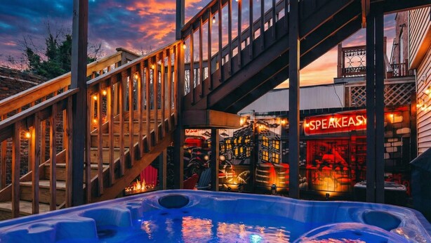 A hot tub area with wooden railings and a view of a building with a neon sign. - *New* Speakeasy Highlands 7BR | Hot Tub | Sleep 14 (Louisville)