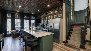 A modern kitchen with a central island, bar stools, and a staircase leading to an upper level. - *New* Speakeasy Highlands 7BR | Hot Tub | Sleep 14 (Louisville)