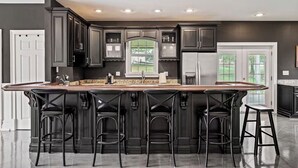 A modern kitchen with dark cabinetry, a central island with bar stools, and a large window providing natural light. - Bourbon Trail Estate | 3 Epic Acres | 7 Bed 6 Bath (Louisville)