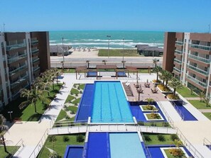 A swimming pool with a view of the ocean, surrounded by palm trees and a building with balconies. - Hosts BR - Apartamentos no VG Fun (Fortaleza)