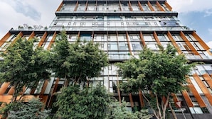 En moderne bygning med flere etager og en glasfacade, omgivet af frodig vegetation og træer.