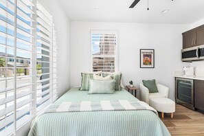 A bedroom with a bed, a chair, a small table, a window with blinds, and a kitchen area with cabinets and appliances.