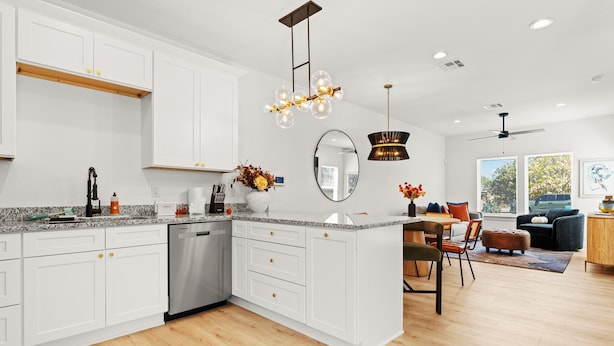 A modern kitchen with white cabinetry, a built-in dishwasher, and a central island with a sink. There is a dining area with a table and chairs, and a ceiling fan. - Sweet September - Stylish Waco Retreat in a Prime Location (Waco)