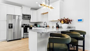 A modern kitchen with white cabinetry, a stainless steel refrigerator, a microwave, and a large island with bar stools. - Sweet September - Stylish Waco Retreat in a Prime Location (Waco)