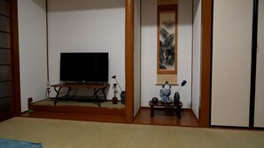 A traditional Japanese room with tatami flooring, a television on a wooden stand, a painting on the wall, and a small table with a vase and a figurine. - GINTAKEI (Osaka)