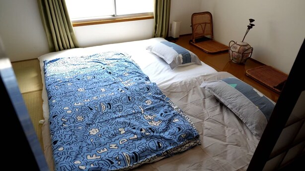 A neatly made bed with a patterned blue blanket and white pillows, a small wicker chair, a decorative vase with dried flowers, and a woven mat on the wooden floor. - GINTAKEI (Osaka)