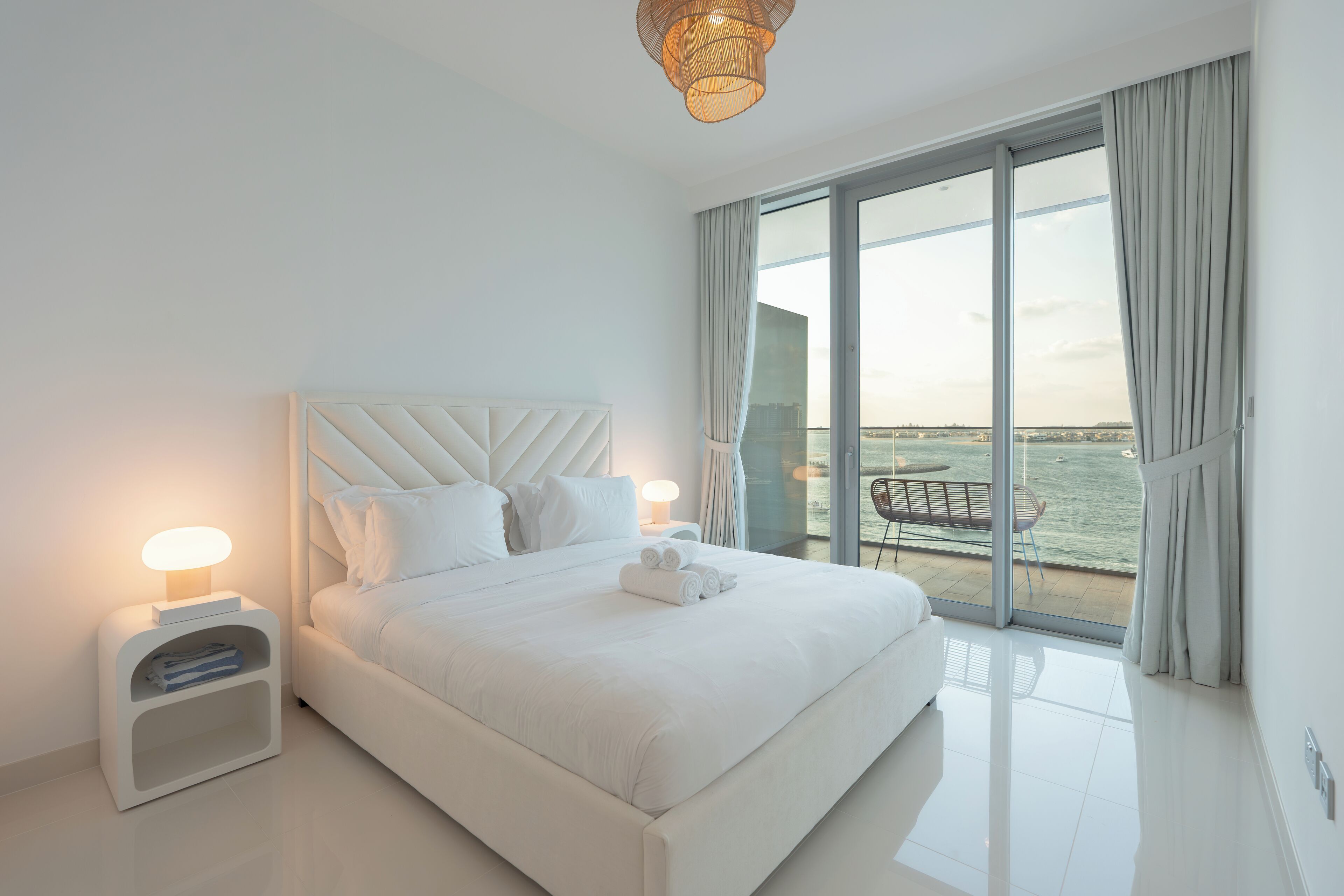 A modern hotel room with a large bed, a bedside table with a lamp, and a view of the sea through floor-to-ceiling windows.