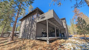 A two-story cabin with a covered porch and a small deck, surrounded by trees and a snowy ground. - Alpine 4 - SHARC, Hot Tub, A/C, Light & Bright! (Sunriver)