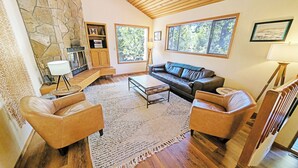 A spacious living room with a stone fireplace, leather sofas, a coffee table, and large windows offering a view of the outdoors. - Alpine 4 - SHARC, Hot Tub, A/C, Light & Bright! (Sunriver)