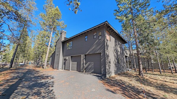 A two-story house with a garage, surrounded by tall pine trees. - Alpine 4 - SHARC, Hot Tub, A/C, Light & Bright! (Sunriver)