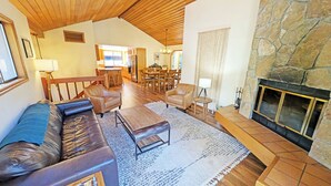 A spacious living room with a fireplace, wooden furniture, and a view into the kitchen. - Alpine 4 - SHARC, Hot Tub, A/C, Light & Bright! (Sunriver)