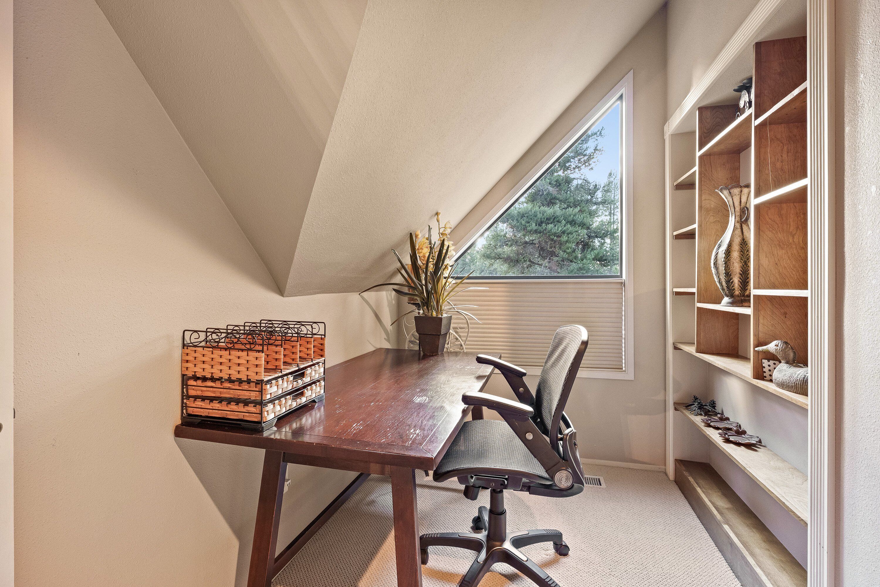 Une pièce avec un plafond en pente, un bureau en bois, une chaise, une plante en pot et une bibliothèque intégrée.