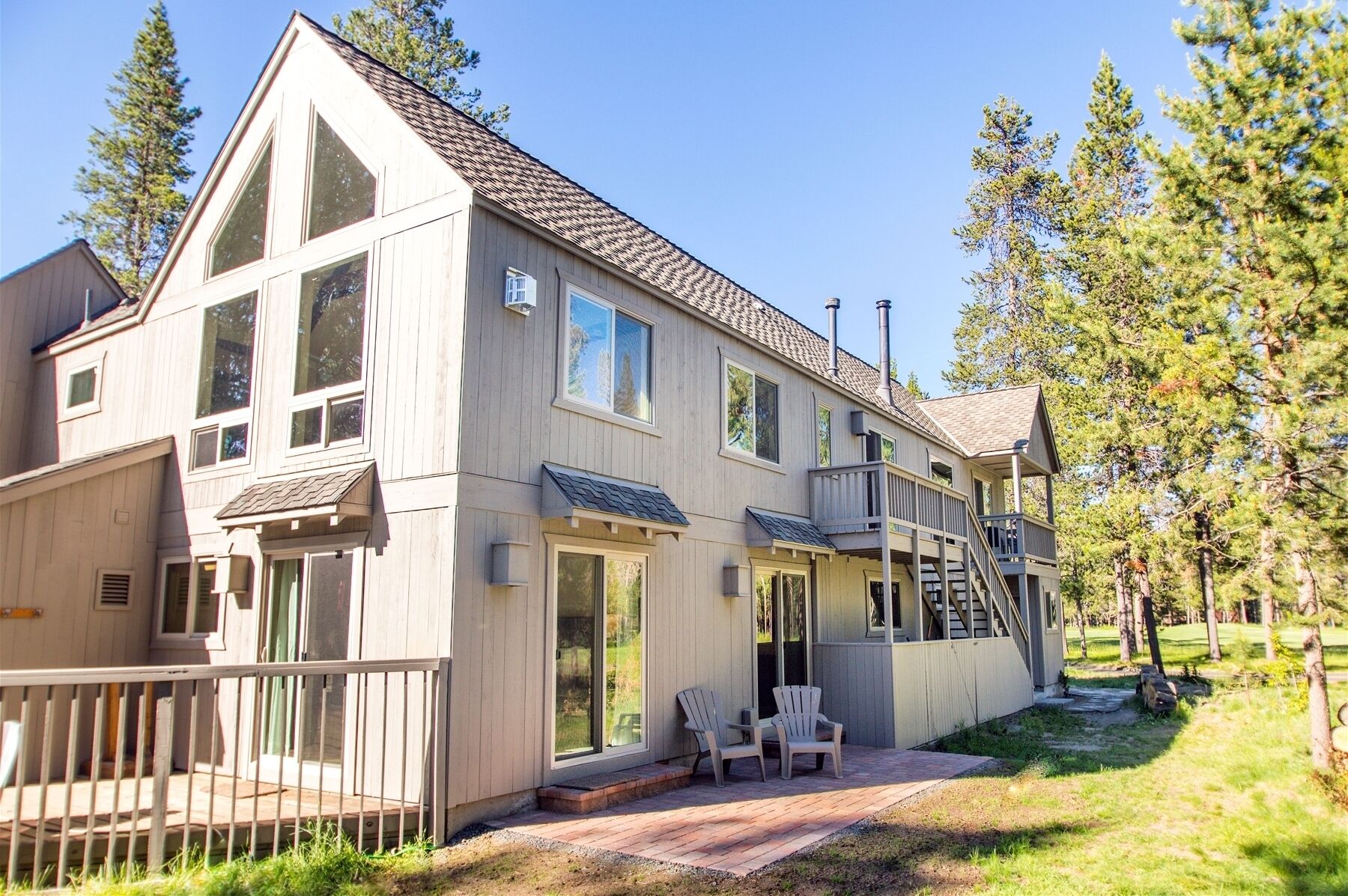 A two-story wooden house with a balcony, large windows, and a small deck area with chairs.