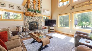 A spacious living room with a stone fireplace, large windows, and a television. - Aspen Place - River Access, Ping Pong, Gorgeous Home & Location (Three Rivers)