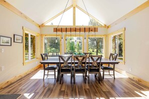 A dining area with a wooden table and chairs, large windows, and a high ceiling with exposed beams.