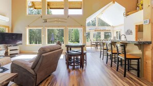 A spacious living area with a large window, a fireplace, and a dining area with wooden chairs and a table. - Aspen Place - River Access, Ping Pong, Gorgeous Home & Location (Three Rivers)