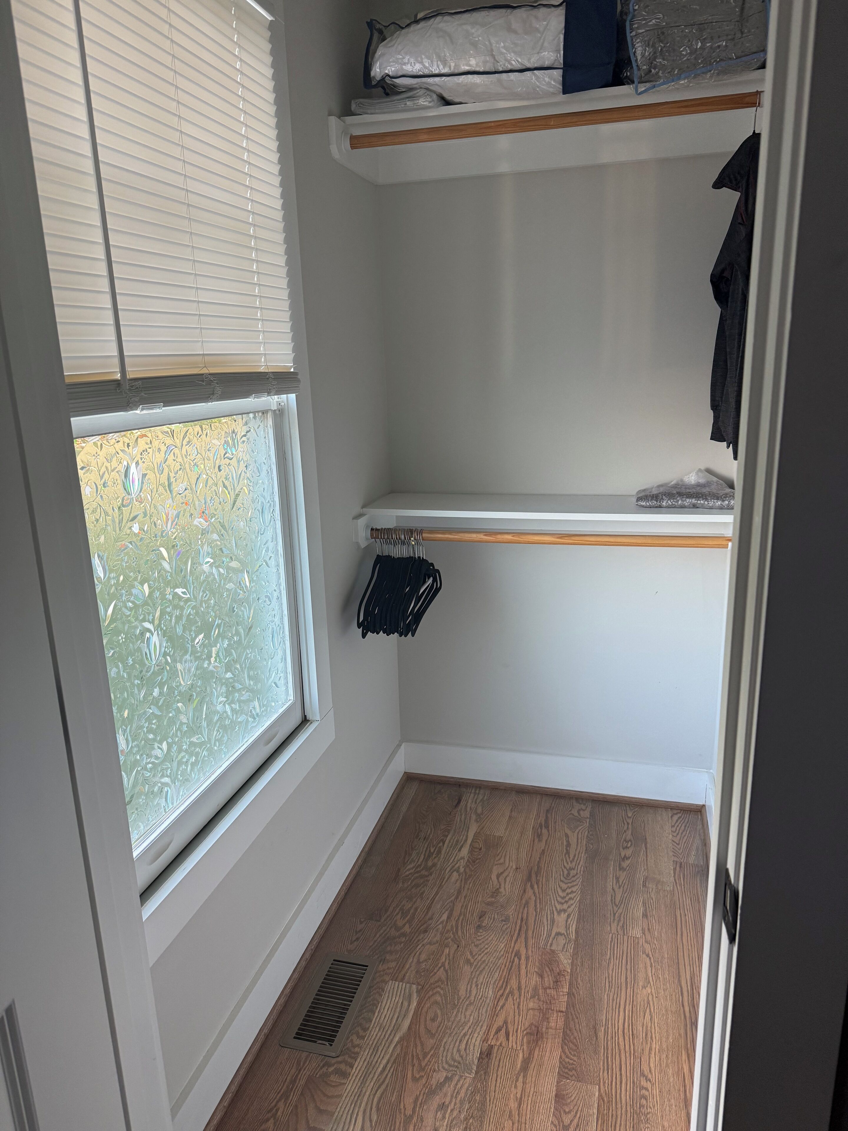 A corner of a room with a window, a wooden floor, a white wall, and a shelf with folded clothes and a bag.