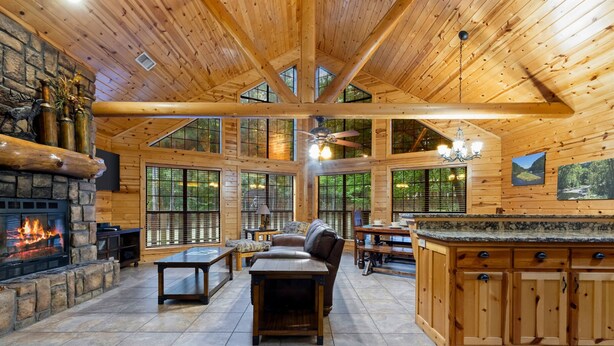 A spacious lodge interior with a stone fireplace, wooden ceiling beams, large windows, and a kitchen area.
