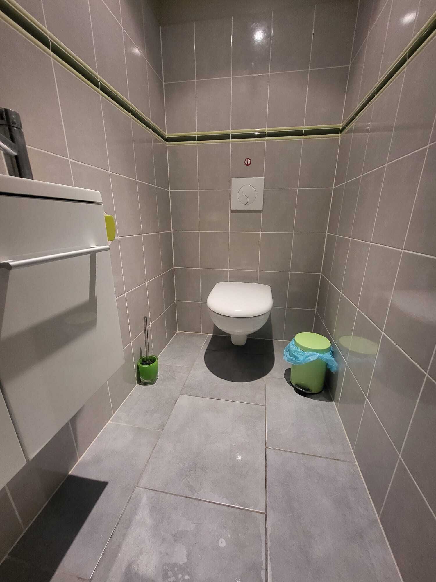 Une petite salle de bain carrelée, avec des toilettes, une poubelle verte et un seau de nettoyage vert.