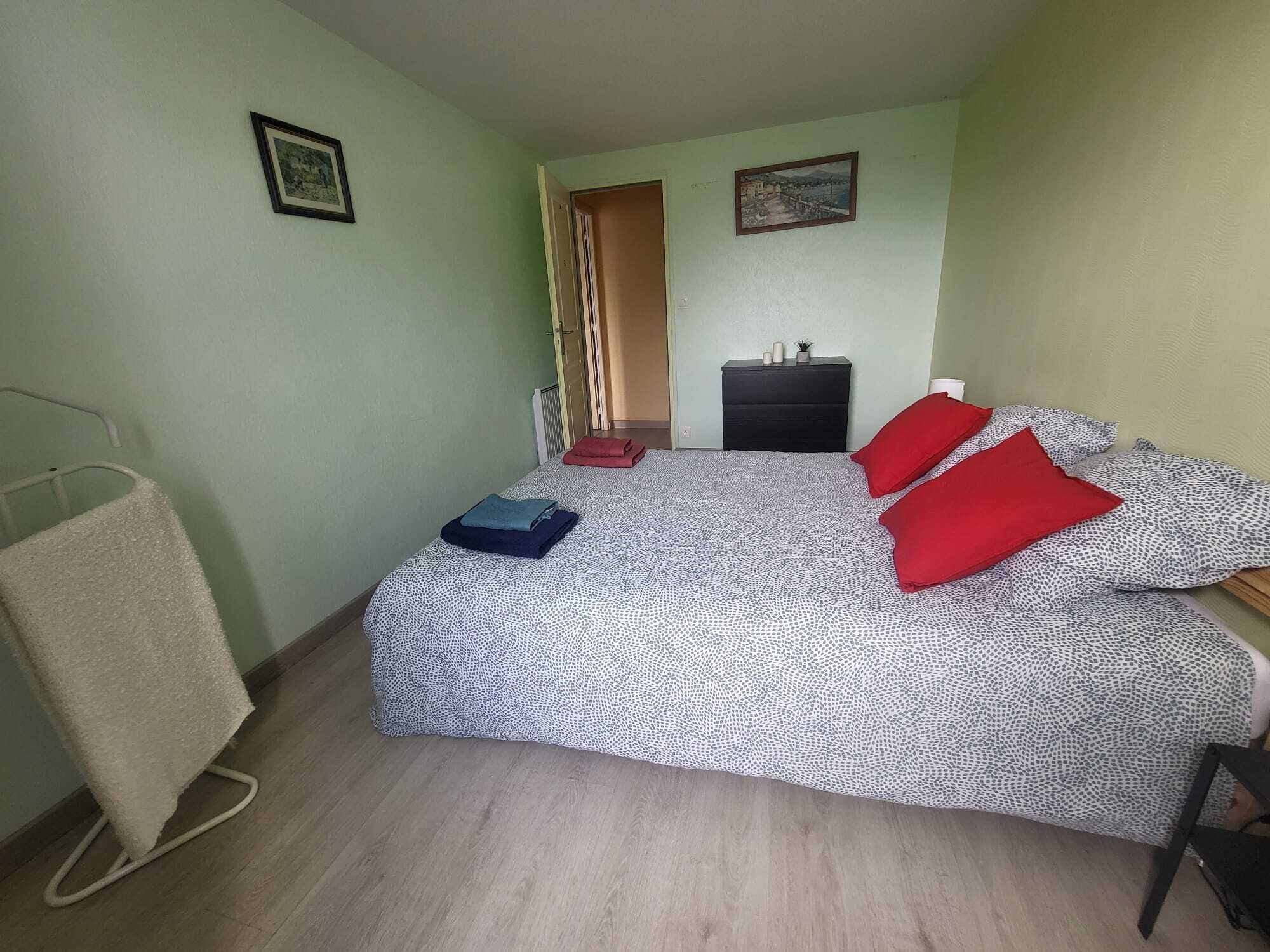 Ein kleiner, ordentlich eingerichteter Schlafzimmer mit Bett, Nachttisch, Stuhl, Heizkörper und einer Tür zu einem anderen Raum.
