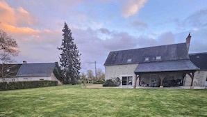 A grassy outdoor area with a stone building featuring a covered patio and a large tree.
