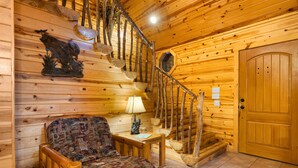 Ein Holzblockhaus-Interieur mit einer Holztreppe, einem gemusterten Sessel, einem kleinen Tisch mit Lampe und einer Holztür.