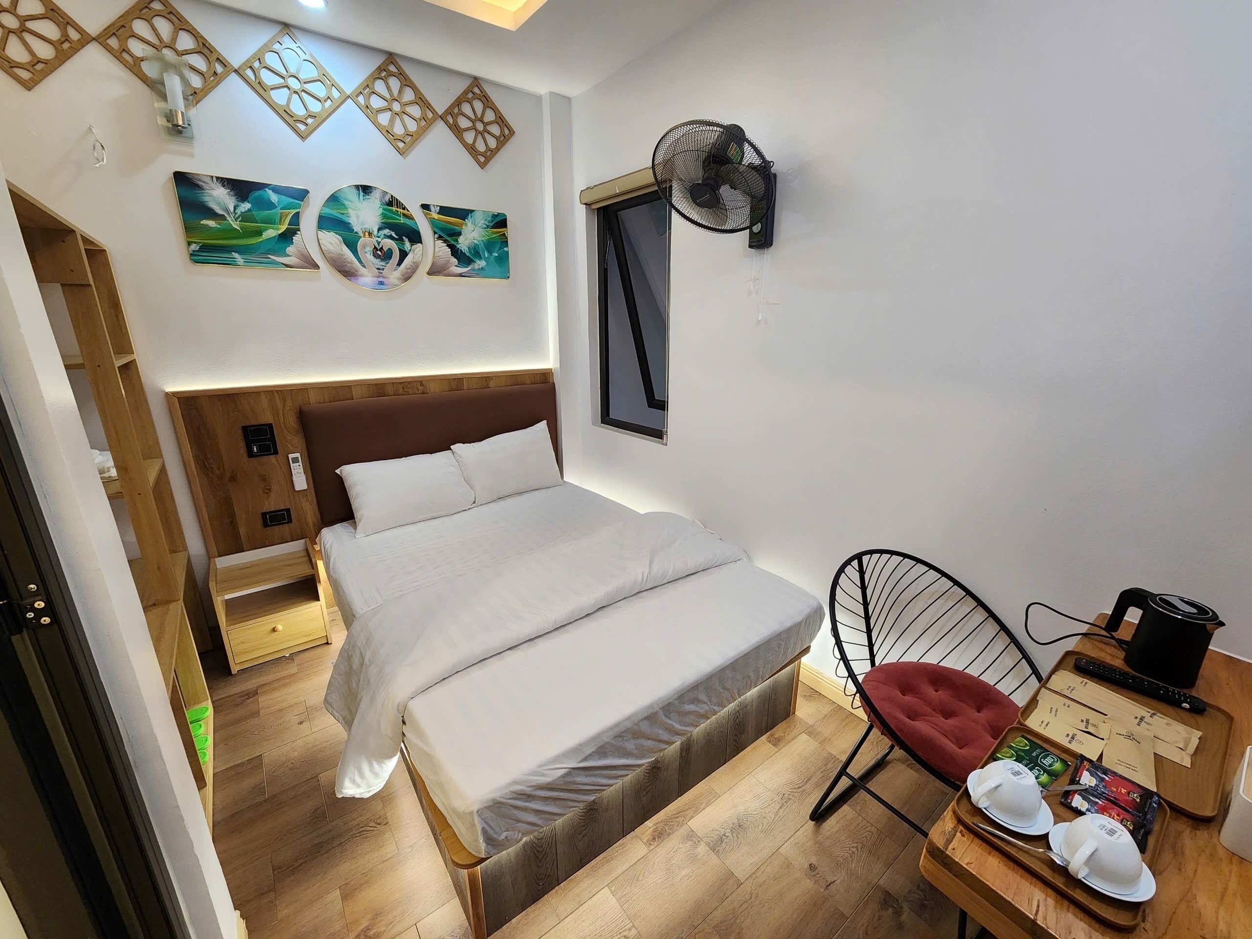 A neatly arranged bedroom with a wooden headboard, a bed with white linens, a wall-mounted fan, and a small table with a tray holding a cup and a remote control.
