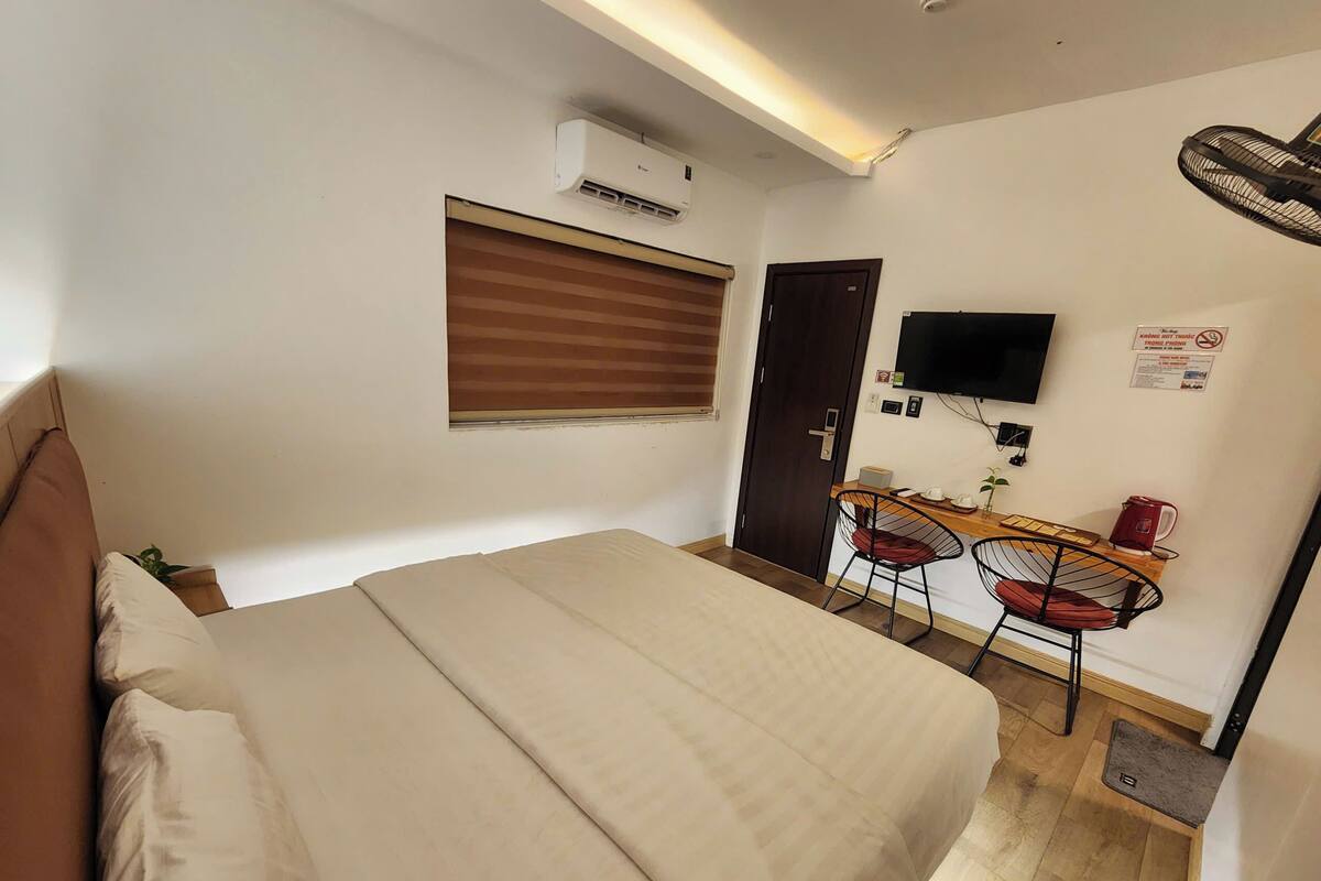 A compact hotel room with a bed, a desk with a chair, a television, and an air conditioning unit.
