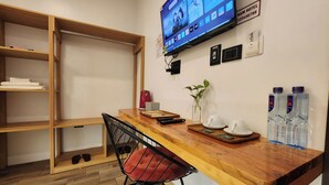 Zona de recepción de un hotel con un mostrador de madera, un televisor montado en la pared, un estante con toallas dobladas, un jarrón con planta y botellas de agua.