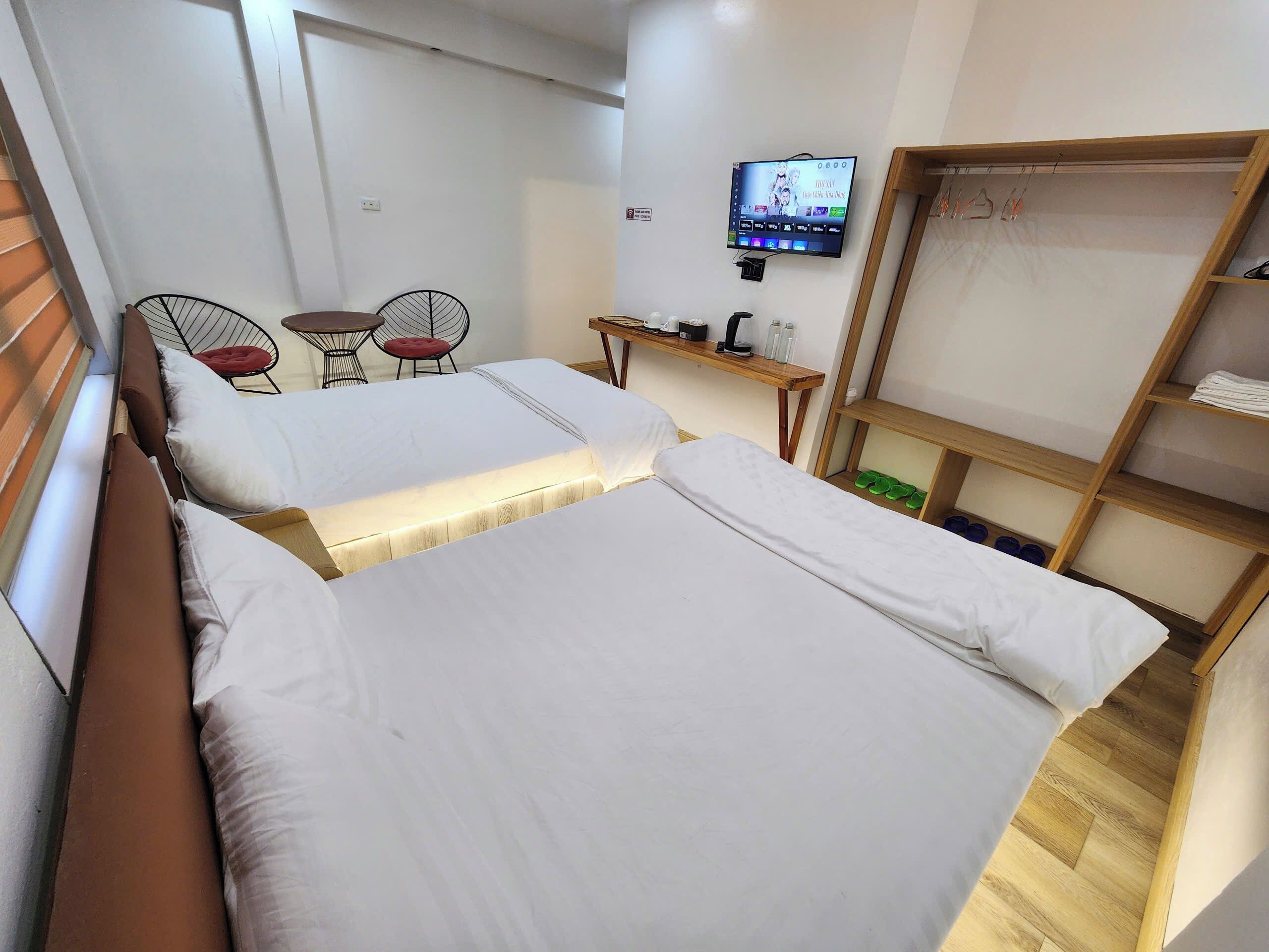 A hotel room with two beds, a TV mounted on the wall, a small table with chairs, and a wooden shelf with shoes.
