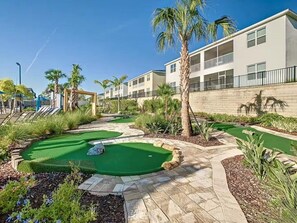 Un campo de golf con múltiples áreas de práctica de putt, palmeras y un edificio con balcones al fondo.