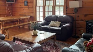 Een knus interieur van een chalet met een groot raam, houten wanden, een leren bank, een bijzettafeltje en een aan de muur bevestigde televisie.
