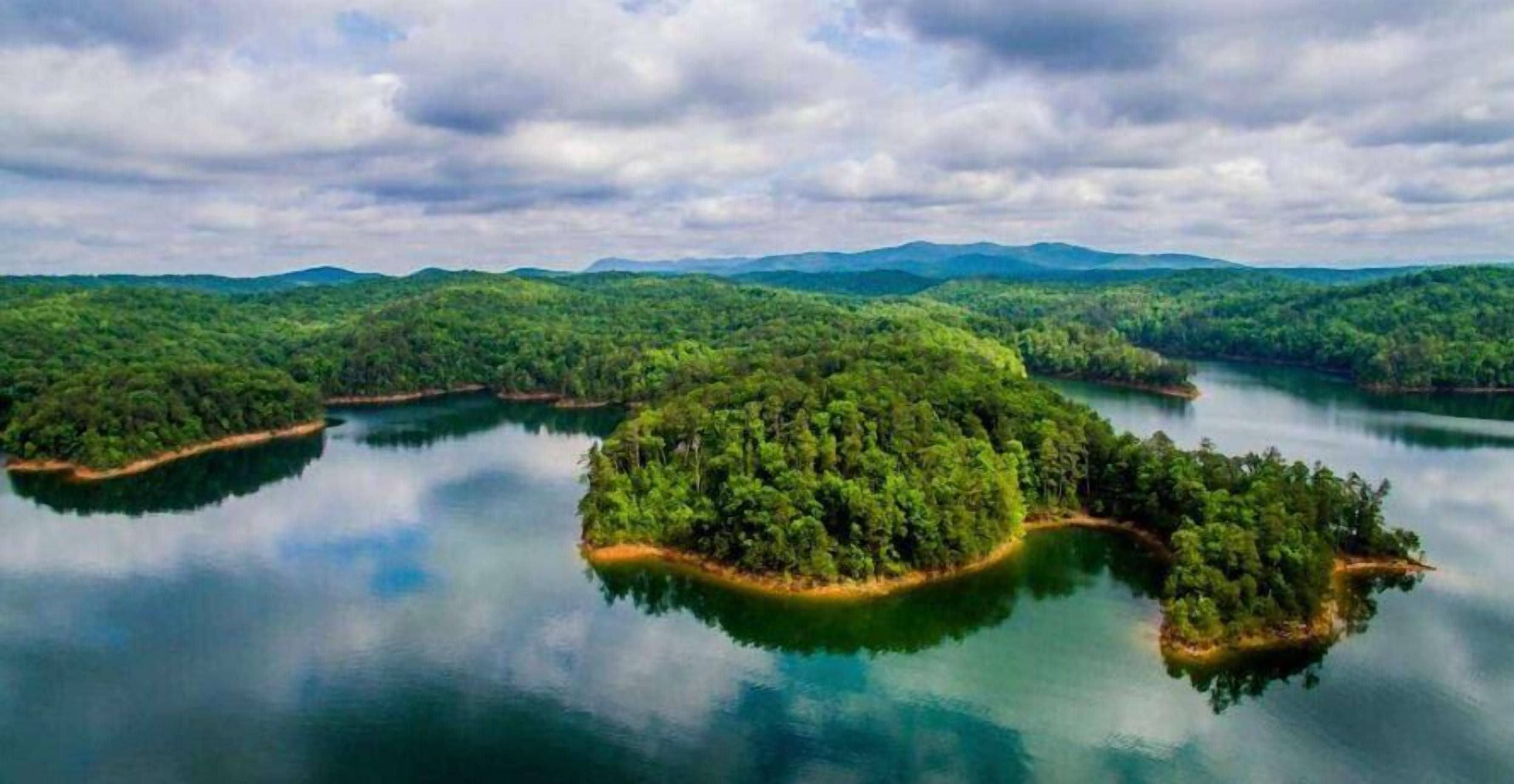 Een rustig landschap met meerdere eilanden omringd door kalm water, weelderige begroeiing en verre bergen onder een bewolkte lucht.