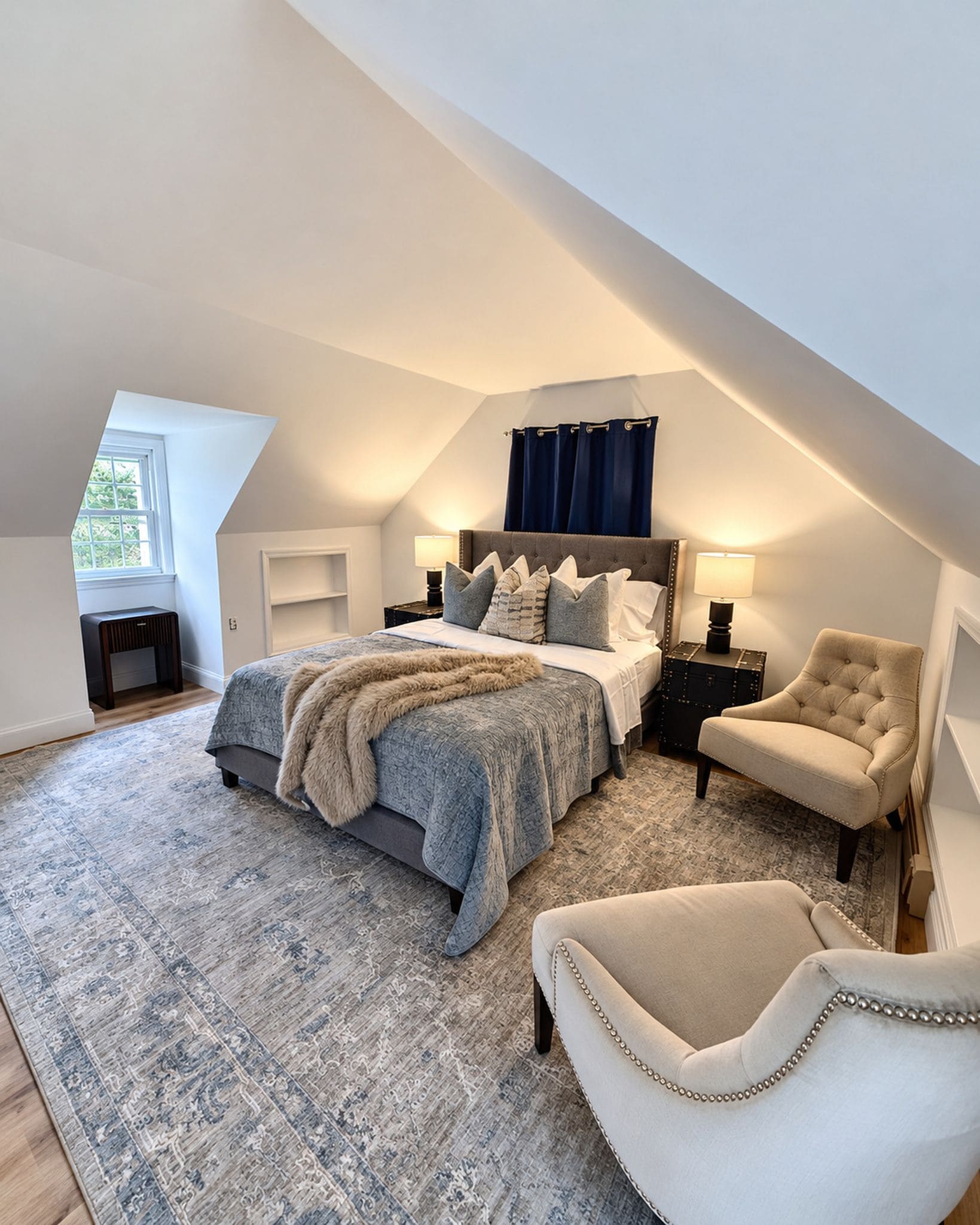 A bedroom with a sloped ceiling, a large bed with a fluffy blanket, two bedside tables with lamps, a chair, and a view of the outdoors through a window.