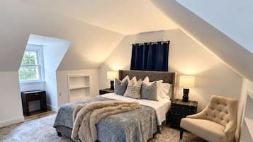 A bedroom with a sloped ceiling, a large bed with a fluffy blanket, two bedside tables with lamps, a chair, and a view of the outdoors through a window.