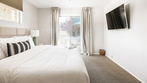 A hotel room with a large bed, a flat-screen TV mounted on the wall, a ceiling fan, and a view of the outdoors through a window with curtains.