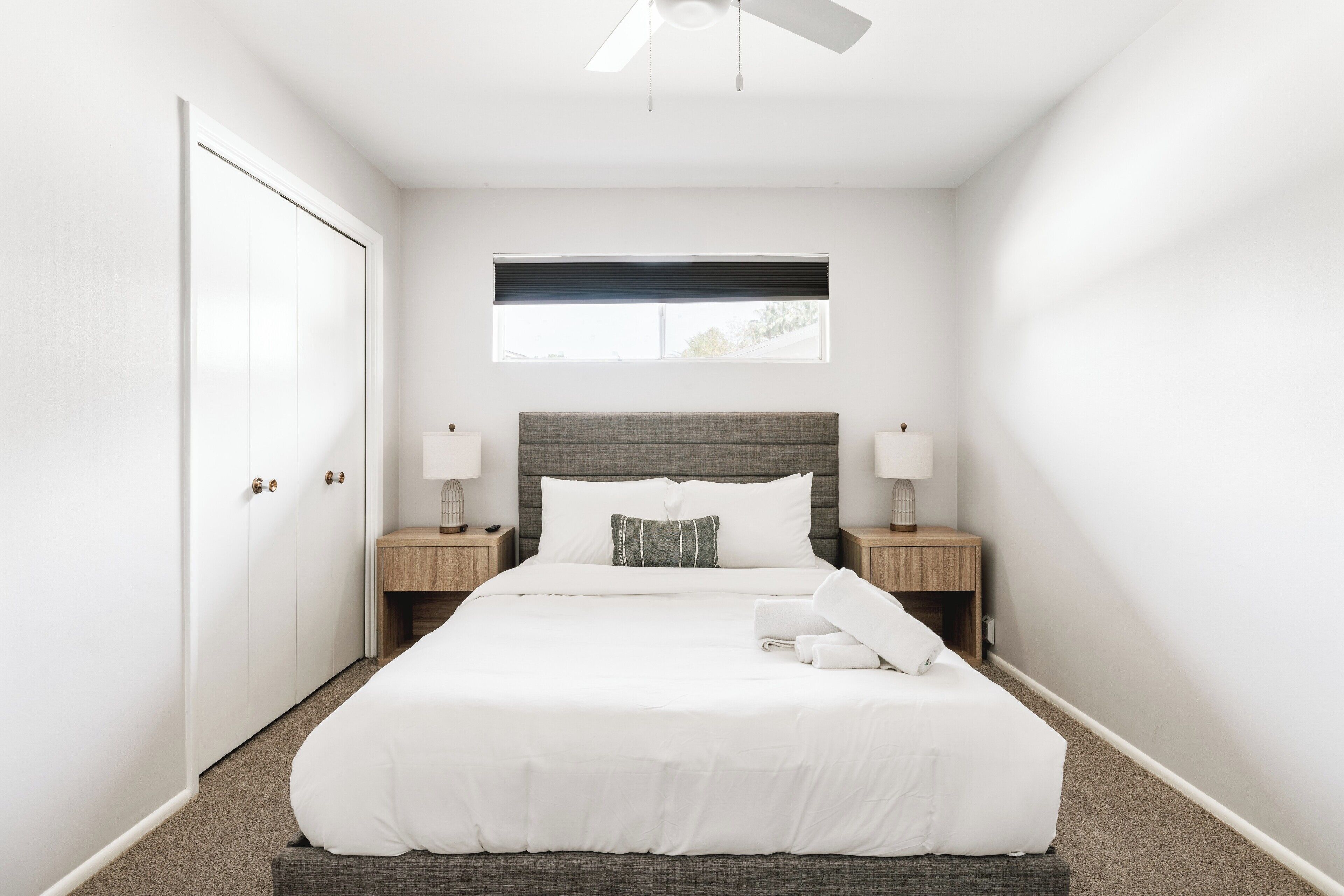 A neatly arranged hotel room with a large bed, bedside tables, a ceiling fan, and a window with blinds.