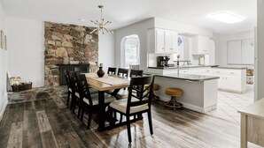 A modern kitchen with white cabinets, a stone accent wall, and a dining area with wooden flooring and black chairs.