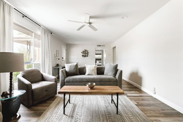 Ein modernes Wohnzimmer mit Sofa, Sessel und Couchtisch, einem Deckenventilator und einem gemusterten Teppich.