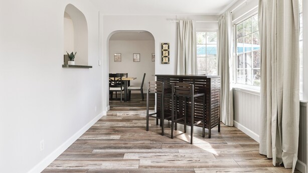 A spacious hallway with a wooden floor, a dining area with chairs and a table, and a window with curtains.
