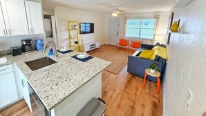 Un apartament modern amb cuina, zona de menjador i saló. Disposa d'un taulell de granat, armaris blancs, un televisor de pantalla plana i un sofà amb coixinets taronja.